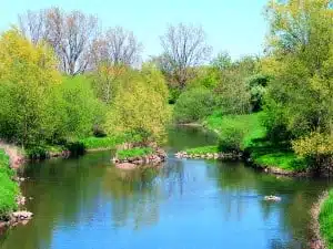 Ein ruhiger Fluss schlängelt sich unter strahlend blauem Himmel durch grüne, grasbewachsene Ufer und Laubwälder – eine perfekte Kulisse für Wanderungen in der Wetterau oder auf den Wanderwegen von Bad Vilbel, wo Felsen das Wasser durchbrechen und üppiges Frühlingslaub gedeiht.