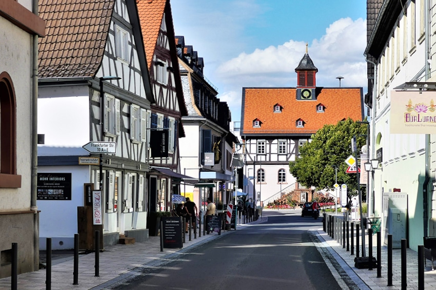 Eine schmale Straße in einer kleinen europäischen Stadt mit Fachwerkhäusern, einem Uhrturm mit rotem Dach am Ende und Menschen, die dort spazieren gehen – das erinnert an die Routen entlang der Wanderwege von Bad Vilbel oder an die malerischen Wanderwege im Rhein-Main-Gebiet an einem sonnigen Tag.