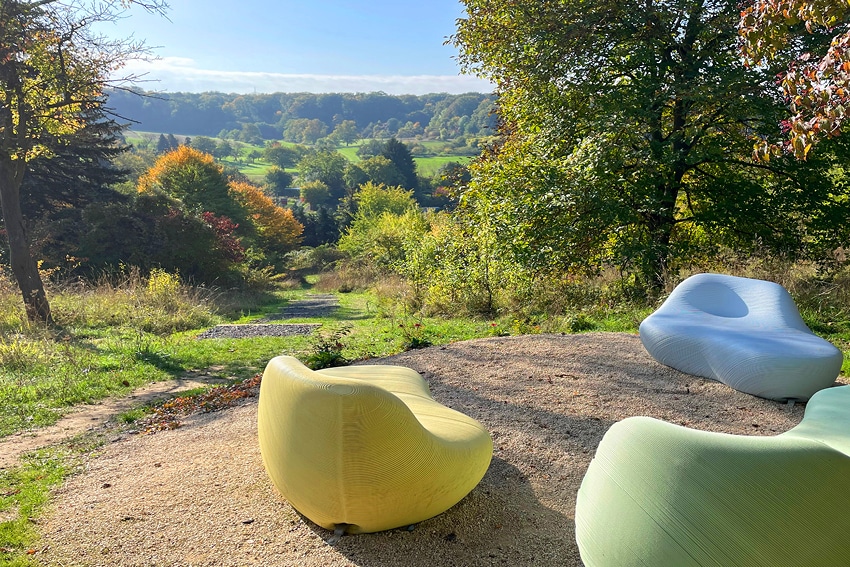 Drei moderne, farbenfrohe Außenbänke stehen auf einer sandigen Fläche neben einem Weg, umgeben von Bäumen und üppigem Grün – ideal für eine Pause, während man die Wanderungen in der Wetterau, das Wandern in der Nähe von Frankfurt oder die Wanderwege von Bad Vilbel genießt.