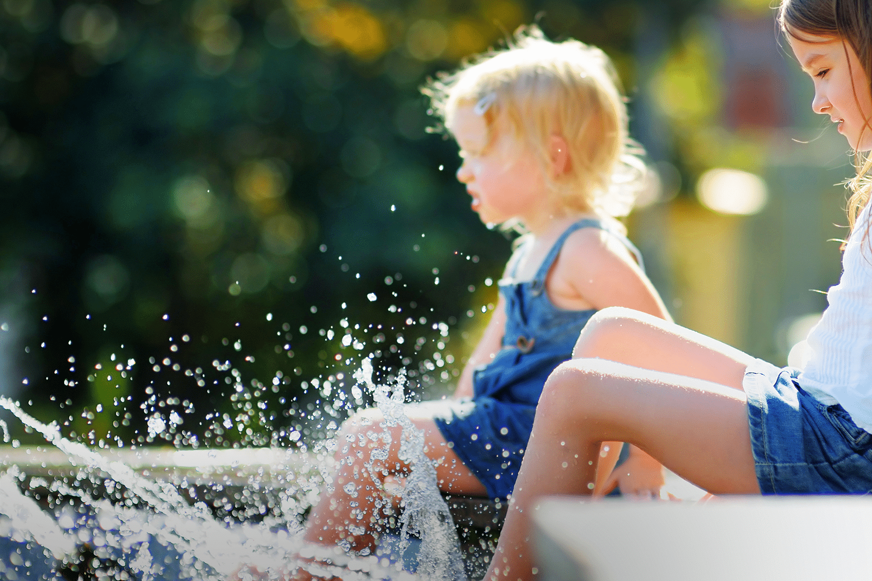 Zwei kleine Kinder sitzen am Rand eines Brunnens und planschen an einem sonnigen Tag mit den Füßen im Wasser – vielleicht machen sie gerade eine Pause vom Erkunden der Wanderwege in Bad Vilbel. Das Sonnenlicht lässt ihr Haar und ihre Gesichter vor einem verschwommenen grünen Hintergrund strahlen.