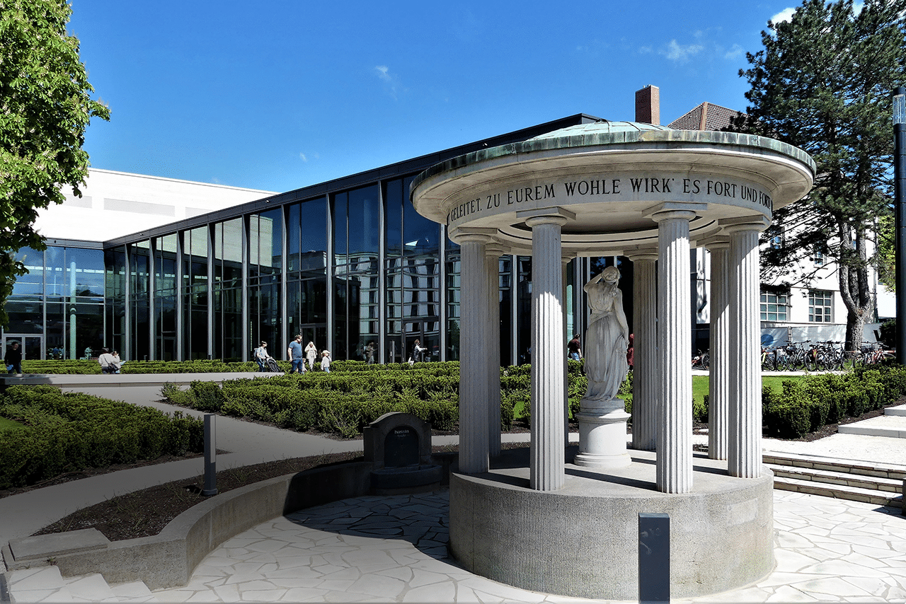 Vor einem modernen Glasgebäude steht ein kleiner, runder Pavillon mit Säulen und einer Statue im Inneren, umgeben von Sträuchern und Bäumen. Er eignet sich perfekt für eine Pause während der Erkundungstouren auf den Wanderwegen von Bad Vilbel; an sonnigen Tagen spazieren die Menschen dort und entspannen sich im Freien.