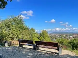 Von zwei leeren Bänken auf einem Schotterweg aus hat man einen malerischen Blick auf eine Stadt – ideal für Wanderfreunde in Bad Vilbel –, mit grünen Bäumen im Vordergrund und einem blauen Himmel mit vereinzelten Wolken darüber.