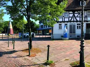 Ein mit Kopfsteinpflaster gepflasterter Platz mit Bäumen, ein Fachwerkhaus, eine große dekorative Flaschenstatue und eine kegelförmige Skulptur im Hintergrund – ideal für alle, die gerne in Bad Vilbel oder in der Wetterau wandern. In der Nähe eines modernen Supermarkts steht ein weißes Auto.