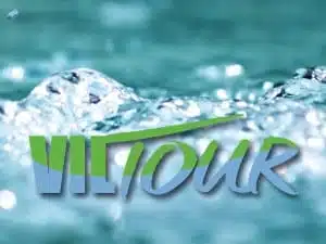Das Bild zeigt das Wort „VITour“ in fetter, stilisierter blau-grüner Schrift vor einem Hintergrund aus spritzendem Wasser, was an das erfrischende Erlebnis der Wanderwege in Bad Vilbel und der malerischen Wanderungen in der Wetterau erinnert.