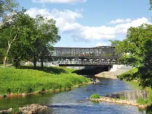 Eine moderne Fußgängerbrücke aus Glas und Metall mit Sitzgelegenheiten im Freien überspannt einen ruhig fließenden Fluss, umgeben von grünen Bäumen und grasbewachsenen Ufern – ideal für alle, die die Wanderwege von Bad Vilbel erkunden oder unter blauem Himmel das Wandern in der Nähe von Frankfurt genießen möchten.