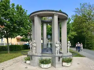 In einem von Grünflächen umgebenen Park entlang der Wanderwege Bad Vilbel steht ein runder Steinbrunnen mit sechs kindlichen Statuen. Vier Menschen spazieren auf einem nahegelegenen gepflasterten Weg, während Blumenkübel den Sockel des Brunnens schmücken.