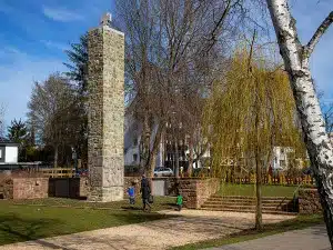 In einem Park entlang der Wanderwege von Bad Vilbel steht ein hohes Steinmonument mit gravierten Tafeln; im Vordergrund sind Bäume, darunter eine Birke, zu sehen, und in der Nähe spazieren an einem sonnigen Tag Menschen, darunter auch Kinder. Im Hintergrund sind Wohnhäuser zu erkennen.