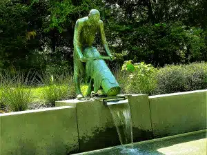 Eine Bronzestatue, die eine sich vorbeugende Person darstellt, die einen großen Fisch in den Händen hält, während Wasser in ein Becken fließt, ist in einem Garten von üppigen Pflanzen und Bäumen umgeben – ein bezaubernder Anblick entlang der Wanderrouten „Wandern Bad Vilbel“ oder „Wandern Rhein-Main“.