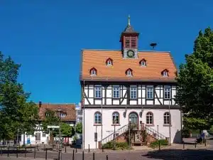 Ein traditionelles Fachwerkhaus mit rotem Ziegeldach, Uhrturm und Außentreppe steht unter strahlend blauem Himmel, umgeben von üppig grünen Bäumen – ideal für Wanderungen in der Wetterau oder zum Wandern in der Nähe von Frankfurt.
