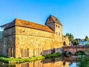 Ein historisches Steingebäude mit einem Obergeschoss in Fachwerk steht an einem ruhigen Fluss – ein idealer Ort zum Wandern in der Nähe von Frankfurt. Eine kleine Bogenbrücke überspannt das Wasser, und unter dem strahlend blauen Himmel versammeln sich die Menschen auf einer Terrasse.