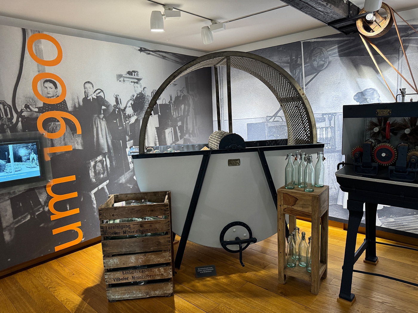 Eine Museumsausstellung zeigt historische Maschinen – wie eine große Trommel, Zahnräder und Kisten mit alten Glasflaschen –, die vor einem Wandgemälde mit Arbeitern und der Aufschrift „um 1900“ zu sehen sind. Ideal für alle, die gerne im Rhein-Main-Gebiet wandern oder kulturelle Touren in der Nähe von Frankfurt unternehmen.