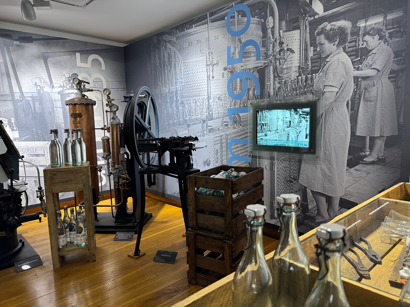 Eine Museumsausstellung zeigt historische Glasflaschen, Holzkisten, alte Maschinen und Werkzeuge. Ein großes Schwarz-Weiß-Foto einer Frau bei der Arbeit in einer Fabrik und ein kleiner Bildschirm erinnern an das industrielle Erbe entlang der Wanderwege von Bad Vilbel.