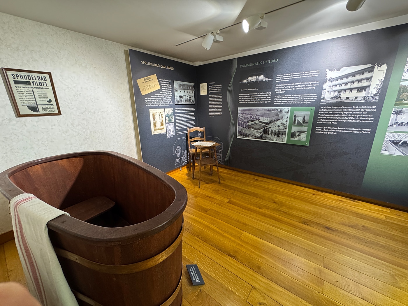 Eine Museumsausstellung mit einer großen Holzbadewanne, einem Stuhl und einem kleinen Tisch versetzt den Besucher in die Geschichte des Kurwesens zurück. An den Wänden sind Fotos und Schautafeln zu sehen, die das kulturelle Erbe der Region beleuchten – darunter auch Routen wie die „Wanderwege Bad Vilbel“. An der Badewanne hängt ein Handtuch; über dem Ganzen leuchten helle Lampen.