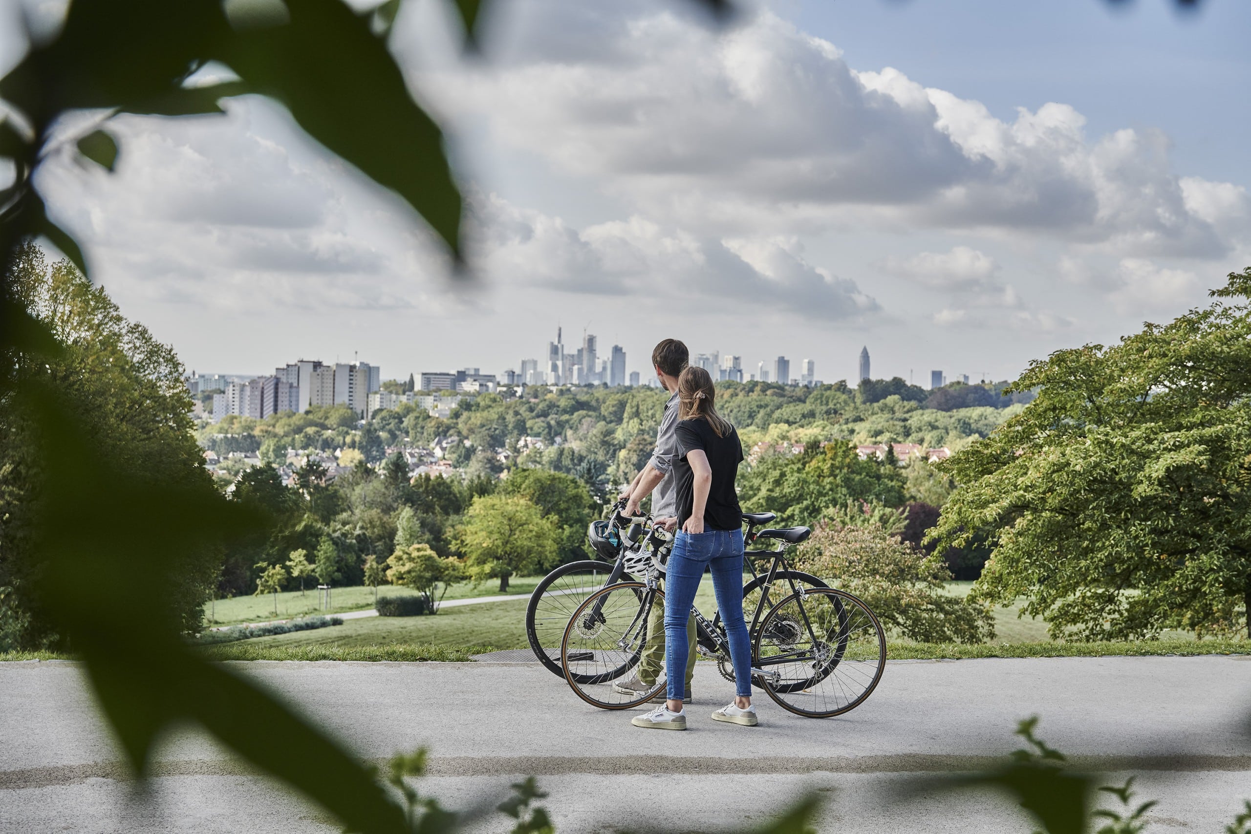 Zwei Menschen mit Fahrrädern stehen auf einem Weg in einem Park und blicken auf die Skyline einer Stadt in der Nähe von Frankfurt, umgeben von Grünflächen und Bäumen unter einem teilweise bewölkten Himmel – ideal zum Wandern in der Nähe von Frankfurt oder zum Erkunden der Wanderwege in Bad Vilbel.