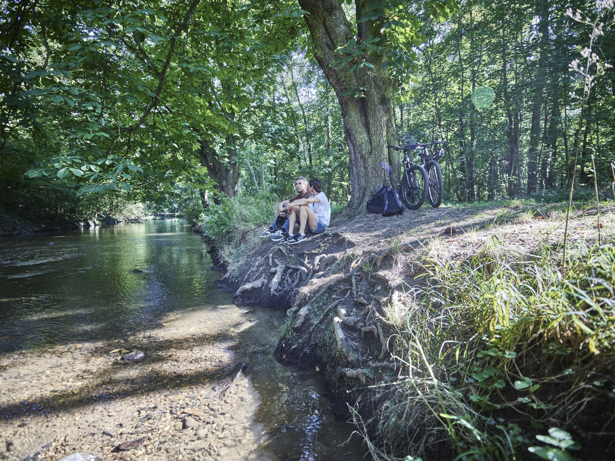 Zwei Menschen sitzen unter Bäumen am grasbewachsenen Flussufer und entspannen sich neben ihren Fahrrädern und Rucksäcken. Das Sonnenlicht scheint durch das üppige Laubwerk entlang der Wanderwege Bad Vilbel, während der ruhige Bach in der Nähe sanft dahinfließt – ideal für Wanderabenteuer im Rhein-Main-Gebiet.