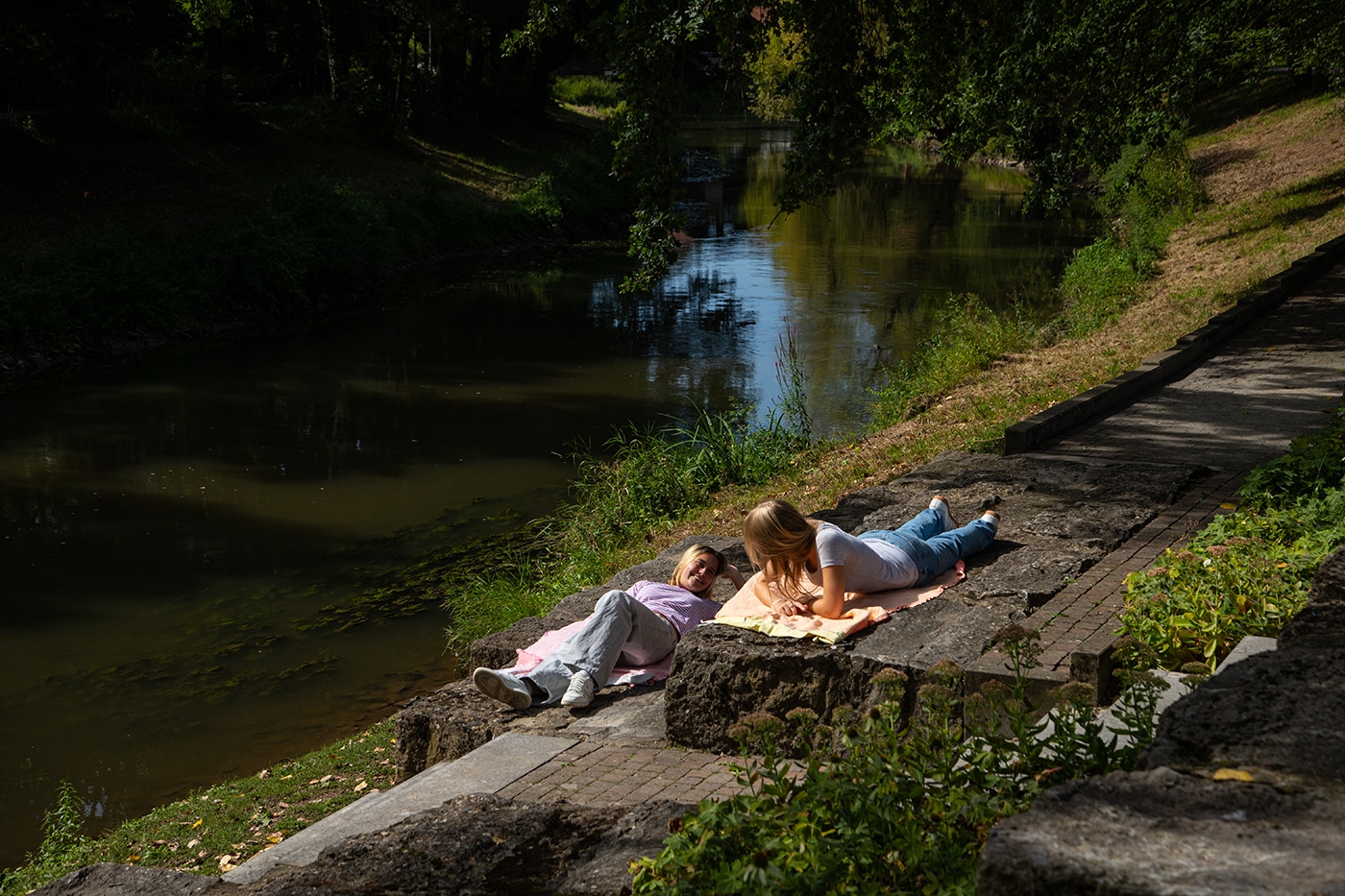 Zwei Menschen liegen auf Decken auf den Steinstufen am Ufer eines ruhigen Flusses und genießen die Sonne und das Grün – ein perfekter Moment, um sich nach einer Erkundungstour auf den Wanderwegen von Bad Vilbel oder der Teilnahme an den Wanderungen in der Wetterau an einem beschaulichen Tag zu entspannen.
