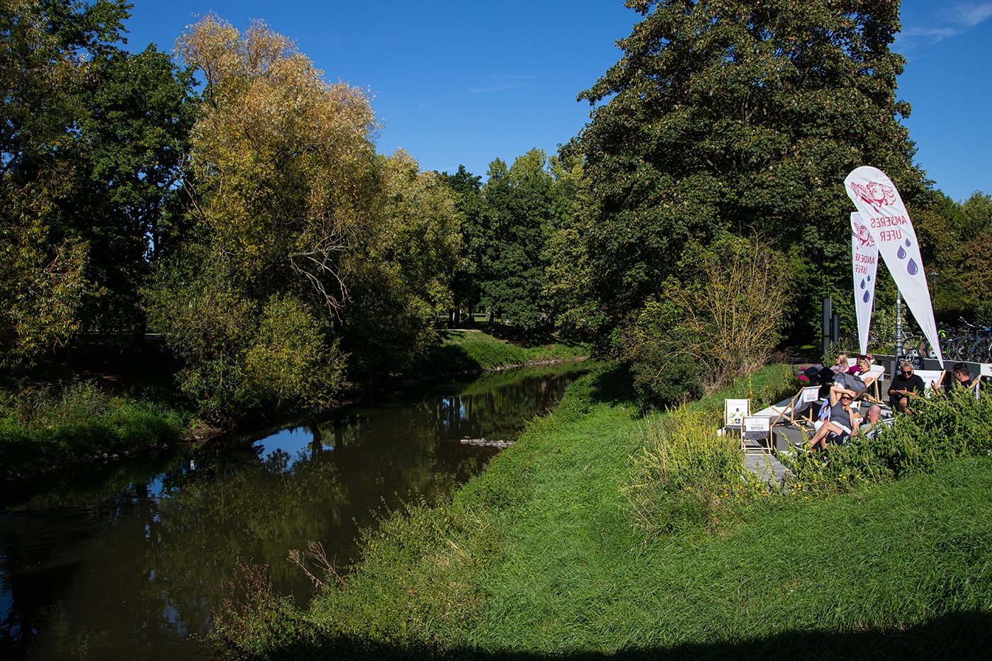 Die Menschen entspannen sich in einem Straßencafé mit Sonnenschirmen in der Nähe eines ruhigen Flusses, umgeben von grünem Gras und hohen Bäumen unter einem strahlend blauen Himmel – ein einladender Ort, nachdem man die Wanderwege Bad Vilbel oder Wanderungen in der Nähe von Frankfurt genossen hat.