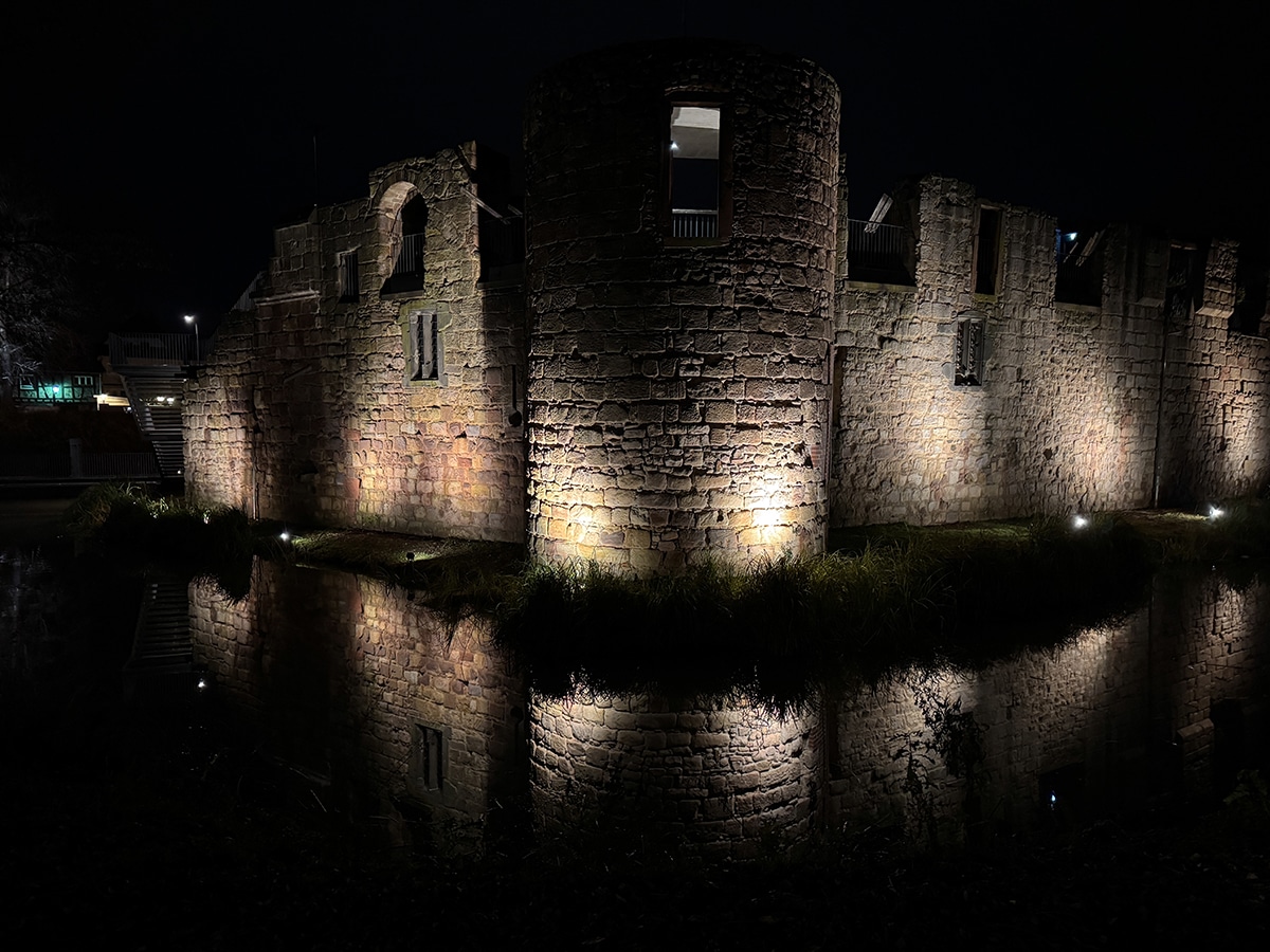Eine alte Steinburg in der Nähe von Frankfurt wird nachts von Bodenleuchten beleuchtet, die dramatische Schatten werfen und sich klar im umliegenden Wasser spiegeln – eine bezaubernde Kulisse für alle, die gerne in der Nähe von Frankfurt wandern.