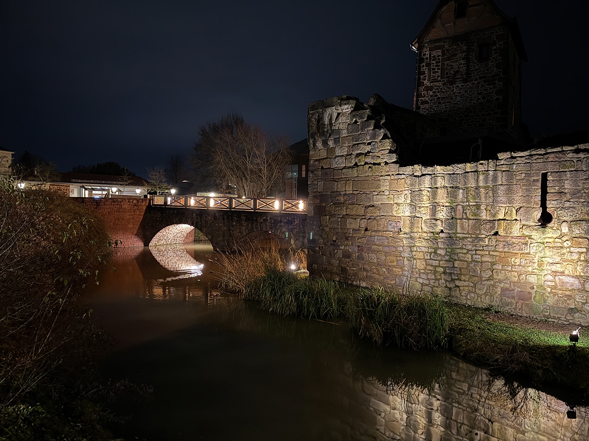 Eine Steinbrücke und eine mittelalterliche Stadtmauer werden nachts beleuchtet und spiegeln sich im ruhigen Wasser darunter wider. Perfekt für „Wandern Rhein-Main“: Unter dem dunklen Himmel säumen vereinzelte Bäume und Gras das Flussufer.