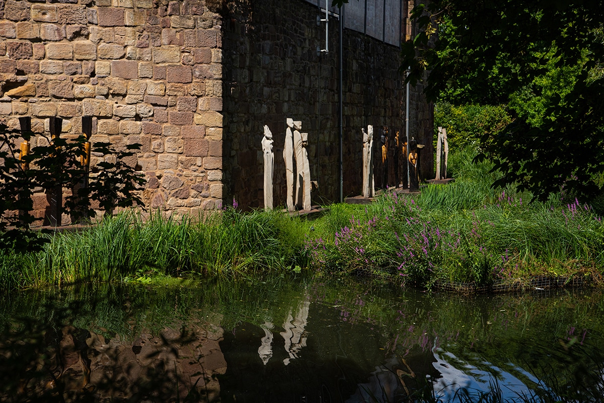 Neben einem Steingebäude reihen sich Holzskulpturen aneinander, umgeben von hohem Gras und Wildblumen – ein einladender Ort für alle, die die Wanderwege von Bad Vilbel genießen, mit schimmernden Spiegelungen im Teich und Sonnenlicht, das durch die nahegelegenen Bäume fällt.