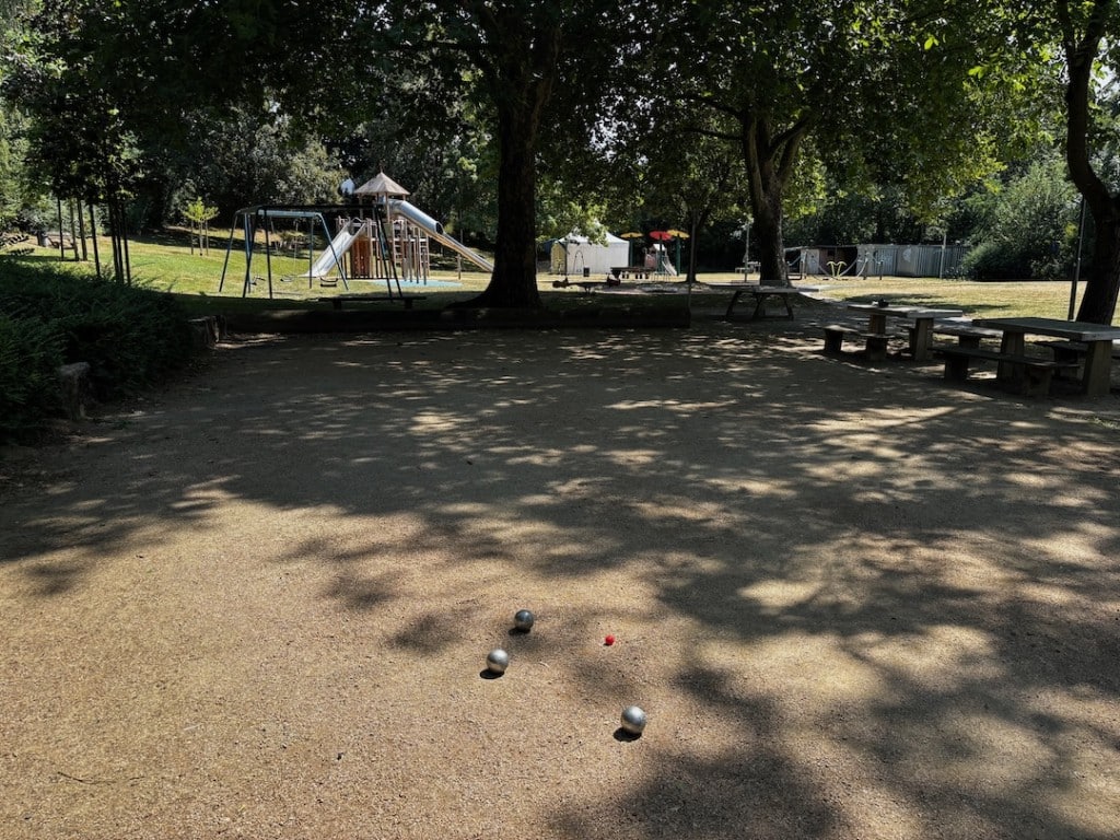 Boules_Massenheim A shady pétanque court with balls and a jack in the foreground. In the background there is a playground with a slide and swings, surrounded by trees.