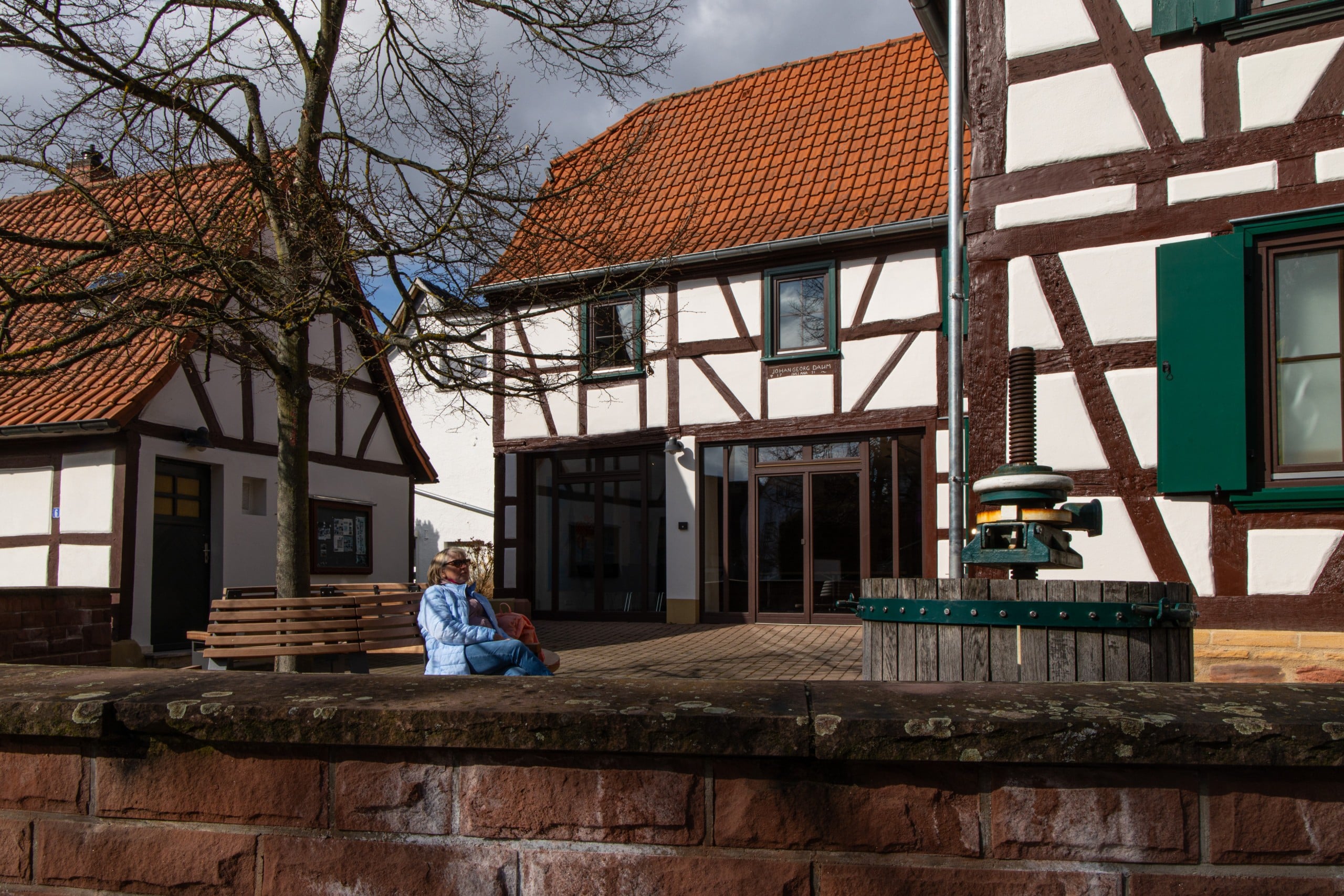 Heimatmuseum Massenheim-1 Eine ältere Frau in einem blauen Mantel sitzt an einem sonnigen Tag auf einer Bank vor traditionellen Fachwerkhäusern mit roten Dächern, unter einem kahlen Baum.