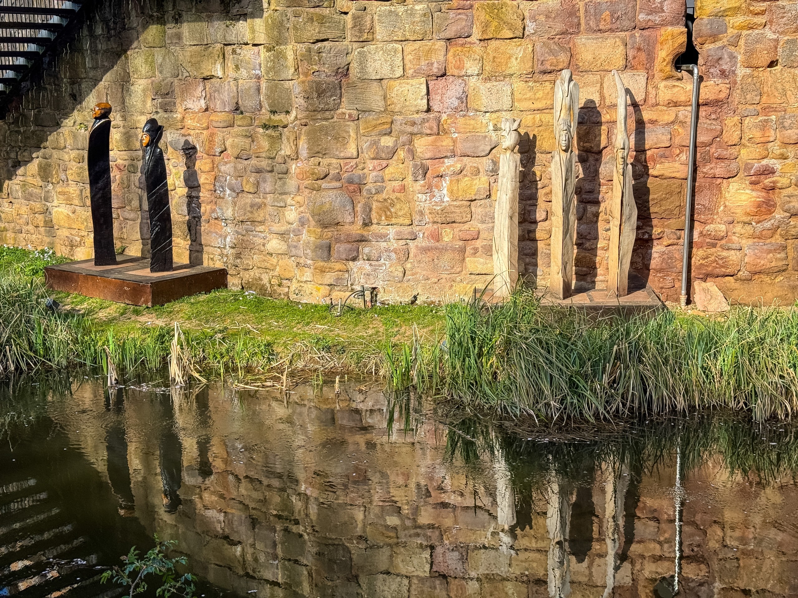Art Guber-1 Four upright wooden sculptures stand beside a stone wall near the water, casting long shadows and reflections. The area is grassy, with some reeds growing along the bank.