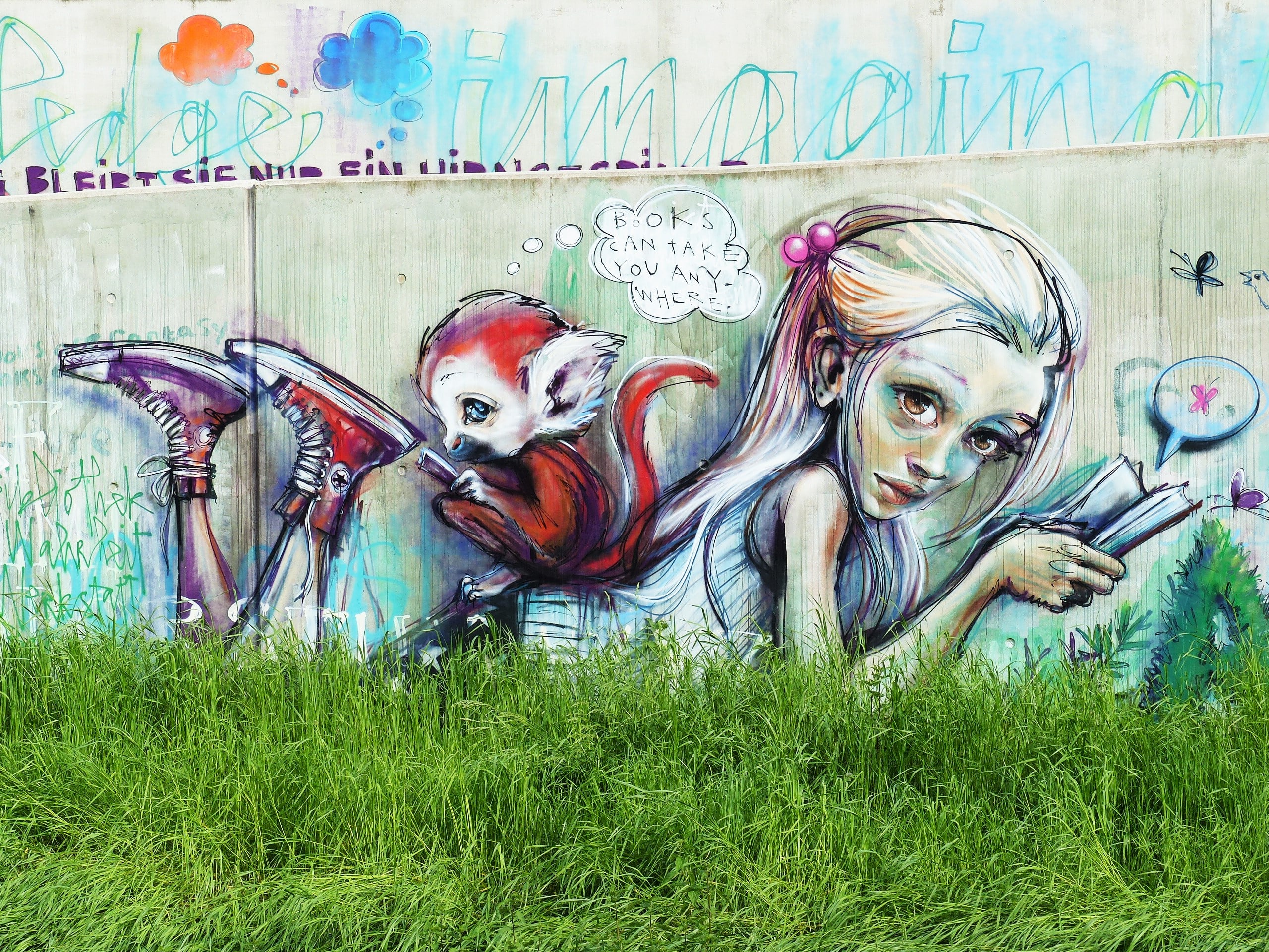 Urban Street Art-1 Street art motif of a girl reading a book, with a small red monkey on her back holding shoes and saying: "Books can take you anywhere." Colorful graffiti is visible in the background, with green grass in front.